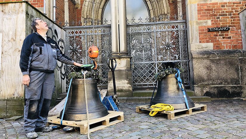 Ein Mann steht neben den beiden neuen Glocken vorm Nordturm und wirft einen prüfenden Blick nach oben. Eine der Glocken ist an einem Seilzug befestigt.  