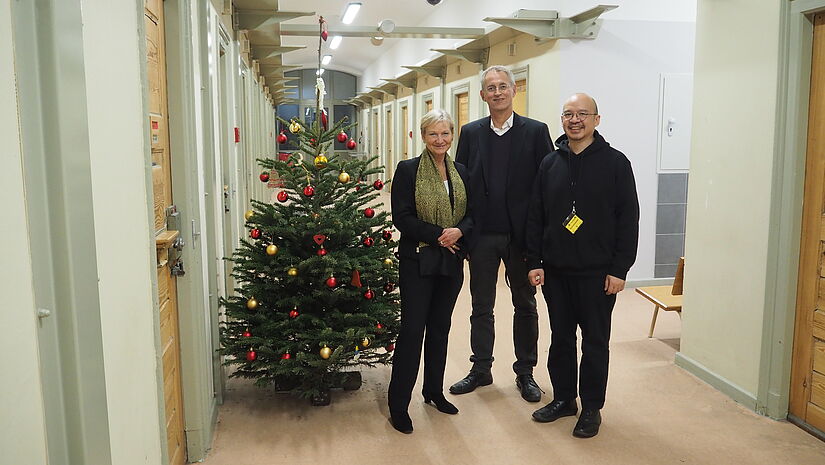 Bischöfin Kirsten Fehrs, Pastor Friedrich Kleine und Kantor Yotin Tiewtrakul (v.l.) stehen im Gefängnis in Hamburg-Fuhlsbüttel vor einem Weihnachtsbaum