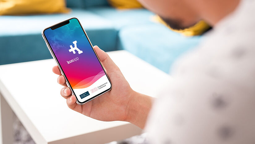 Man schaut einer Person über die Schulter, die ein Smartphone hält. Auf dem Display ist der Startbildschirm der Konapp in blau-pink zu sehen.