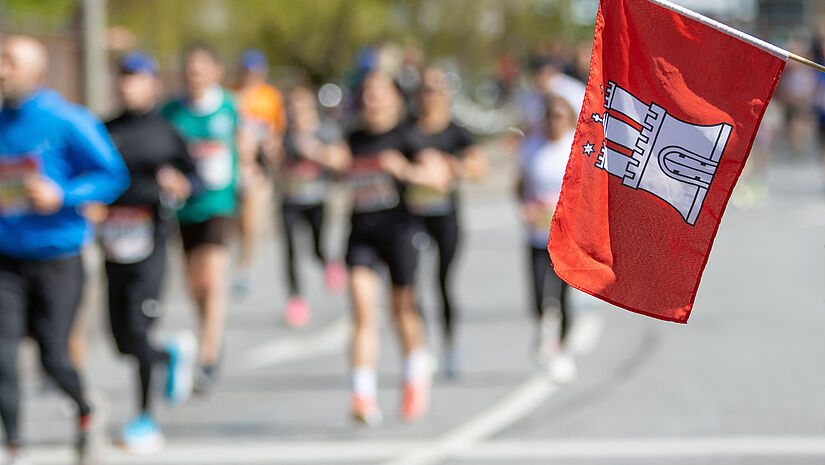 Menschen laufen in Sportkleidung eine Straße entlang. Im Vordergrund schwenkt jemand eine rote Flagge mit dem Hamburg-Wappen.