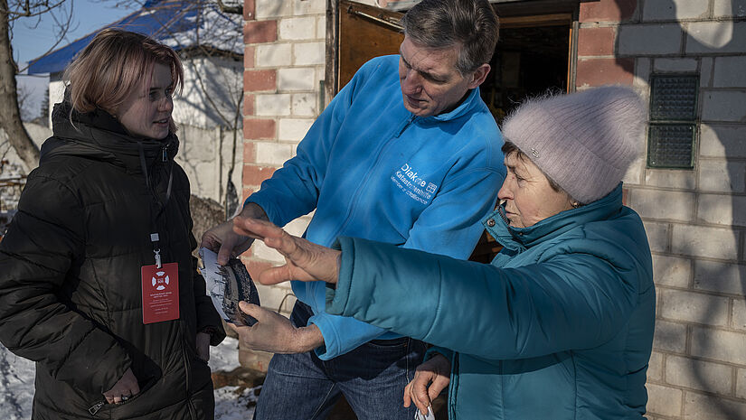 Olena Dyka (rechts) zeigt Martin Keßler, Direktor der Diakonie Katastrophenhilfe, Fotos ihres zerstörten Hauses, das zu Beginn des Krieges 2022 in Ivanivka, etwa zwei Autostunden nördlich der Hauptstadt Kiew, von vorrückenden russischen Truppen zerstört wurde. 