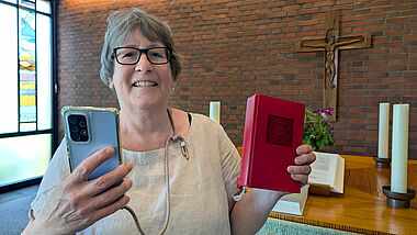 Gudrun Bruhs ist Küsterin der Kirchengemeinde Holm in Schleswig-Holstein. Hier steht sie in der Kirche, in einer Hand hält sie ihr Handy, in der anderen ein Gesangbuch.