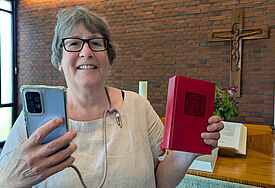 Gudrun Bruhs ist Küsterin der Kirchengemeinde Holm in Schleswig-Holstein. Hier steht sie in der Kirche, in einer Hand hält sie ihr Handy, in der anderen ein Gesangbuch.