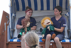 Angelina Thalau (links) und Kristin Schlegel erzählen mit Tom und Tina eine Geschichte im Strandkorb der Tourismuskirche.