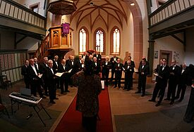 Ein Kirchenchor steht in einer kleinen Kirche, davor die Chorleiterin