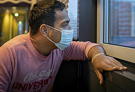 Ein Matrose mit dunklen kurzen Haaren blickt aus einem Fenster der Jugendherberge in Horn. Er trägt einen blauen Mundschutz.
