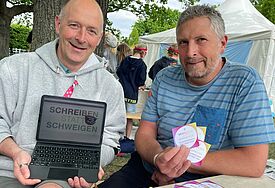 Zwei Männer halten ein IPad und Aufkleber mit dem Schriftzug Schweigen statt Schweigen in ihren Händen.