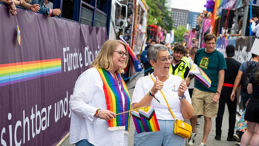 Zwei Frauen winken mit Regenbogenflaggen vor dem CSD-Truck der Kirche beim CSD in Hamburg 2026
