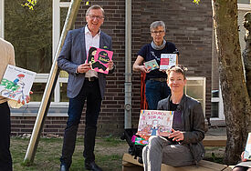Fünf Frauen und Männer stehen und sitzen mit Abstand zueinander vor einer Hauswand. Sie halten Bücher für Kinder vor sich. 