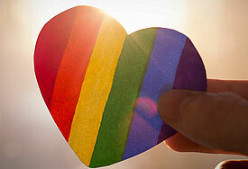Sonne scheint von hinten auf ein Herz aus Papier aus Regenbogenfarben gehalten von einer Hand.