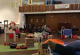 Vor der Orgel im Kirchengebäude sitzt eine Gruppe Kinder. Die Kirchenbänke sind ausgeräumt, es gibt viel Platz und Spielzeug für die Kleinen.