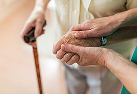 Zwei Hände einer Pflegerin halten die Hand einer älteren Patientin. In der anderen Hand hält sie den Griff eines Gehstocks.