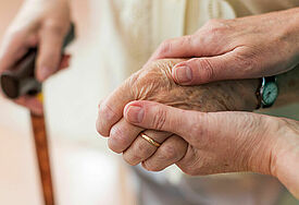 Zwei Hände einer Pflegerin halten die Hand einer älteren Patientin. In der anderen Hand hält sie den Griff eines Gehstocks.