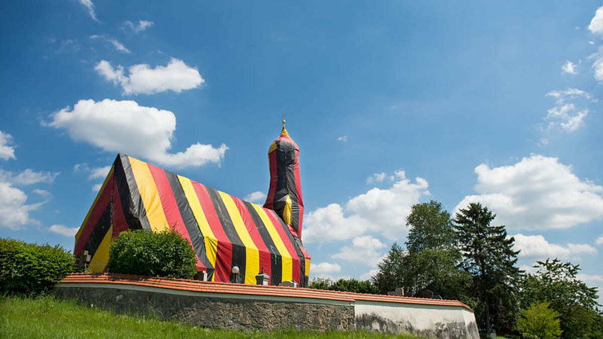 Wuerde der Fussballgott ein passendes Zuhause suchen - im oberbayerischen Kirchseeon koennte er fuendig werden (Foto vom07.07.2016). Passend zur Europameisterschaft in Frankreich ist die Kirche St. Coloman (Foto) in der Gemeinde Kirchseeon seit einigen Tagen in Schwarz-Rot-Gold gehuellt. Doch mit Fussball hat die Aktion nichts zu tun - vielmehr steckt der Wurm drin. Grund fuer die Komplett-Verhuellung des kleinen Gotteshauses sei nicht die EM, sondern eine Holzwurmbekaempfung, sagte der katholische Pfarrer Georg Menachery am Donnerstag (07.07.2016) dem Evangelischen Pressedienst (epd). (Siehe epd-Meldung vom 07.07.2016)