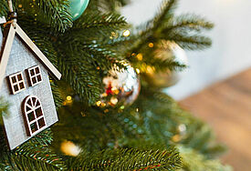 Ein Anhänger aus Holz in Form eines Wohnhauses hängt an einem geschmückten Tannenbaum.