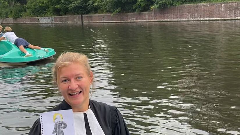 Pastorin Claudia Kress wurde spontan aus einem Boot heraus portraitiert. Sie hält ihr auf Papier gemaltes Bild auf dem SUP stehend vor sich. 