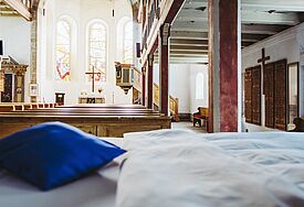 Heller Kircheninnenraum, im Vordergrund steht ein Bett mit weißer Decke und blauem Kissen