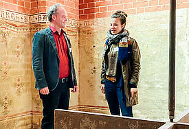 Bischof Tilman Jeremias und Anna Luise Klafs vor dem Kunstwerk „Stigma“, ein an einem langen Pendel herabhängendes Kreuz.