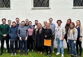 Eine Gruppe von Menschen steht in einer Reihe vor einer hellen Kirchenmauer. 