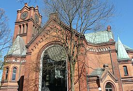 Kirche in Terrakottastein, grüne Dächer, umgeben von zart begrünten Bäumen, der Himmel ist blau, die Sonne scheint