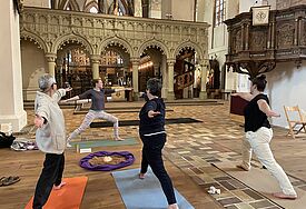 Vier  Frauen praktiizieren die Yoga-Übung "Krieger 1" iin einem leergeräumten Kirchengebäude. 