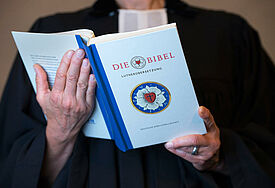 Auf der Bibel ist eine Lutherrose, das Symbol der evangelisch-lutherischen Kirchen. Sie war das Siegel, das Martin Luther ab 1530 für seinen Briefverkehr verwendete.
