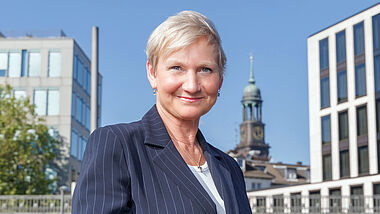 Bischöfin Kirsten Fehrs steht in ziviler Kleidung im Freien, im Hintergrund ist der Turm des Michel zu sehen