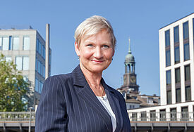 Bischöfin Kirsten Fehrs steht in ziviler Kleidung im Freien, im Hintergrund ist der Turm des Michel zu sehen