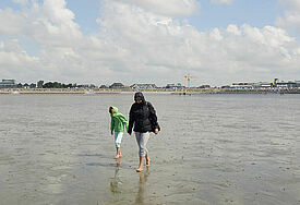 Eine Frau und ihr Kind wandern in Regenjacken durch das Wattenmeer. Im Hintergrund ist der Ort Büsum zu sehen.