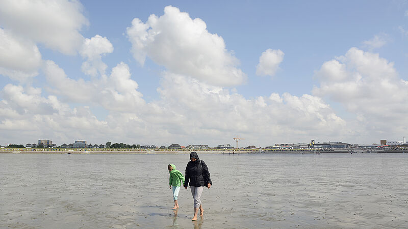 Eine Frau und ihr Kind wandern in Regenjacken durch das Wattenmeer. Im Hintergrund ist der Ort Büsum zu sehen.
