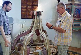 Zwei Männer stehen um einen reich verzierten Schalldeckel im Kircheninnenraum