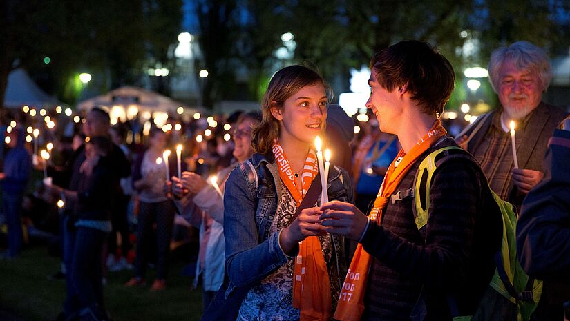 Zwei junge Menschen halten in der Menschenmenge der Messe draußen im Dunkeln zwei Kerzen und lächeln sich an.