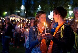 Zwei junge Menschen halten in der Menschenmenge der Messe draußen im Dunkeln zwei Kerzen und lächeln sich an.