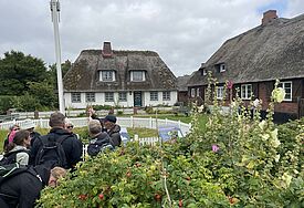 Eine Gruppe Menschen neben zwei Häusern auf Hallig Oland neben dem Wasserspeicherbecken mit Blumene im Vordergrund, 