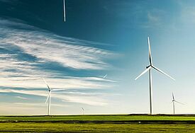 Windräder stehen in einer weiten Landschaft. Am blauen Himmel sind die Kondenzstreifen zweier Flugzeuge zu sehen.