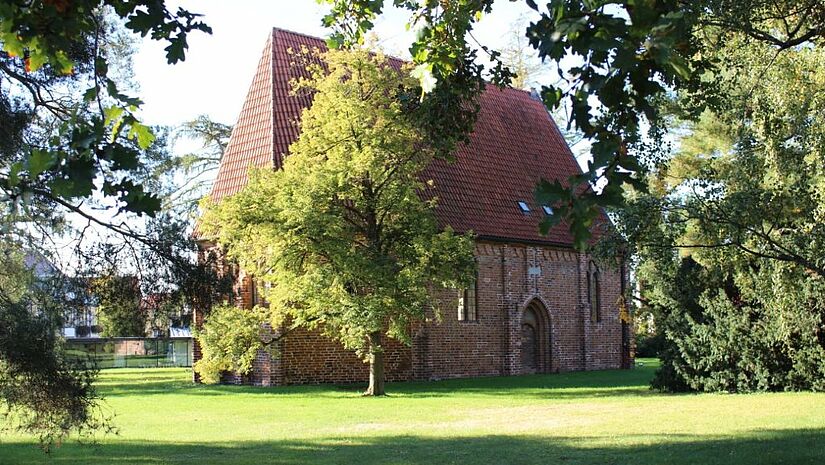 Eine kleine Kapelle steht zwischen grün belaubten Bäumen auf einer Wiese.