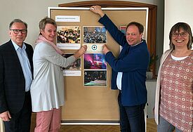 Vier Personen vor einer Stellwand, zwei hängen daran Bilder von Kirchenräumen auf, alle schauen in die KAMERA