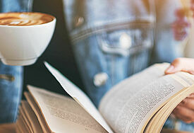 Eine Frau blättert mit einer Hand in einem vor ihr aufgeschlagenen Buch, in der anderen Hand hält sie eine Tasse Kaffee.