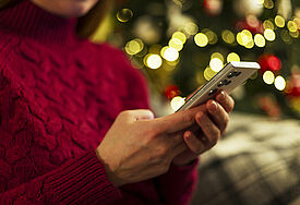 Frau hält ein Smartphone, während sie in der Nähe eines mit Lichtern geschmückten Weihnachtsbaumes sitzt und einen roten Pullover trägt. 