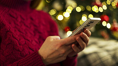 Frau hält ein Smartphone, während sie in der Nähe eines mit Lichtern geschmückten Weihnachtsbaumes sitzt und einen roten Pullover trägt. 