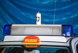 Eine brennende Kerze steht auf einem Autodach mit Blaulicht, im Vordergrund steht auf einem orangen Schild "Notfallseelsorge". 