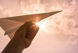 Hand hält einen Papierflieger in die Luft, Sonnenlicht von hinten
