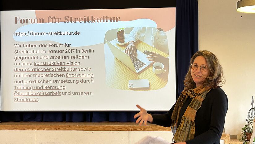 Mareike Brombacher Workshop Mit Andersdenkenren reden lernen Mareike Brombacher steht im Saal und erläutert die Inhalte vom großen Monitor