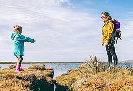 Eine Frau steht mit ihren zwei Kindern am Meer, eins trägt sie auf ihrem Rücken.