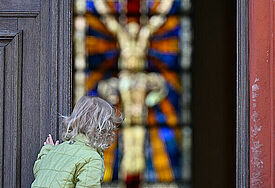 Ein kleines Mädchen schaut vorsichtig durch eine geöffnete Kirchentür. Im Kirchenraum ist ein Kirchenfenster mit Jesus am Kreuz zu sehen.