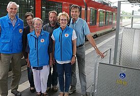 Gruppe an Menschen in blauen Bahnhofsmission-Westen neben einem roten Zug