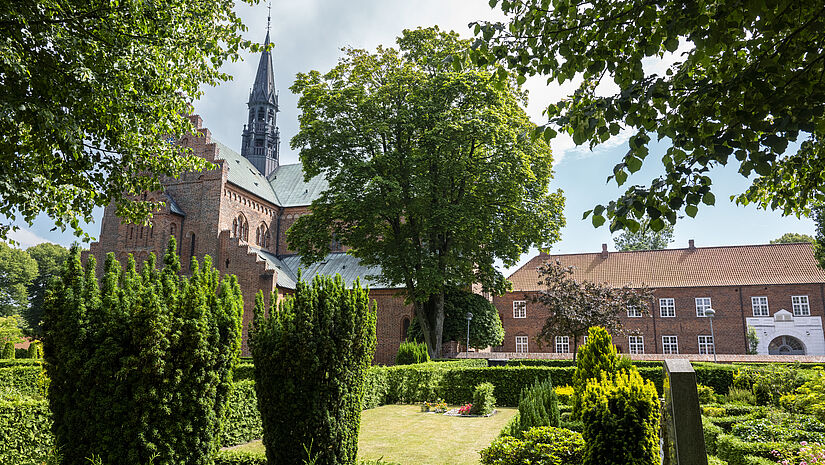 Eine rote Backsteinkirche im gotischen Stil umgeben von grünen Bäumen. 