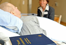 Vorn im Bild liegt das Neue Testament, im Hintergrund unscharf sitzt eine Frau neben einem Krankenhausbett, in dem ein Mann liegt