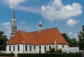 Weiß verputzte Kirche mit roten Dachziegeln und kleinem Turm, umgeben von Laubbäumen