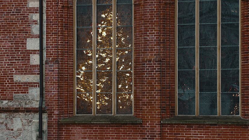 Ein Weihnachtsbaum leuchtet im innern einer Kirche hinter den Fenstern.
